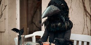 Eine menschengroße Krähe sitzt an einem gedeckten Frühstückstisch, vor ihr Kaffee und ein Ei, an dem eine kleine Krähe pickt