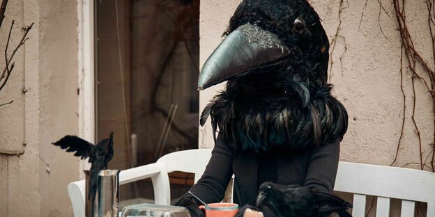 Eine menschengroße Krähe sitzt an einem gedeckten Frühstückstisch, vor ihr Kaffee und ein Ei, an dem eine kleine Krähe pickt