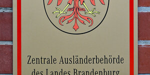 Das Foto zeigt das Schild am Eingang der Zentralen Ausländerbehrde in Eisenhüttenstadt mit dem Namen der Behörde unter dem brandenburgischen Wappen, dem roten Adler.