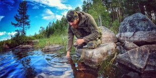 Fritz Meinecke steht an einem Fluss, in den er eine Hand hält, hinter ihm Bäume, neben ihm Felsen