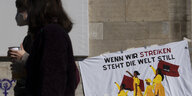 Zwei Frauen laufen an einem Banner vorbei. Anläßich des Frauentages haben sich am 8. März Hunderte Menschen an einer Kundgebung eines Bündnisses aus feministischen Gruppen, Gewerkschaften, Klima- und Care-Aktivisten am Rosa-Luxemburg-Platz versammelt.
