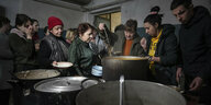 Eine Frau schöpft aus einem riesigen Topf Suppe, um sie herum stehen viele Menschen, die mit Tellern darauf warten, dass ihnen aufgefüllt wird