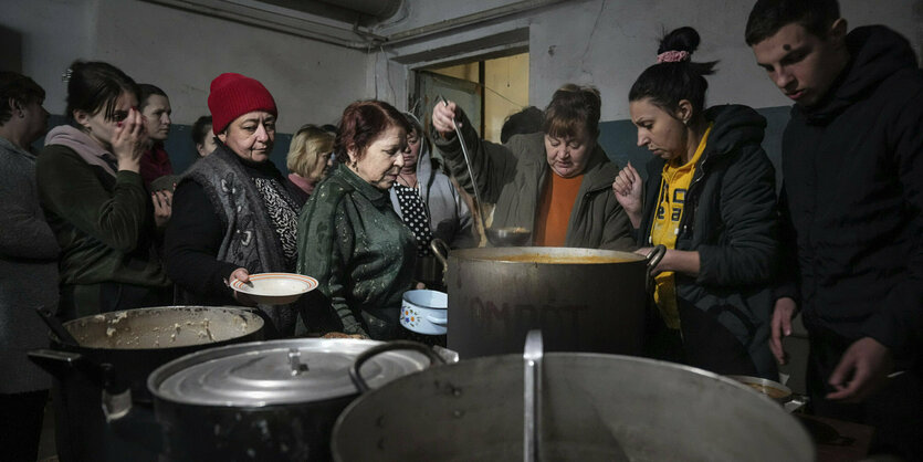 Eine Frau schöpft aus einem riesigen Topf Suppe, um sie herum stehen viele Menschen, die mit Tellern darauf warten, dass ihnen aufgefüllt wird