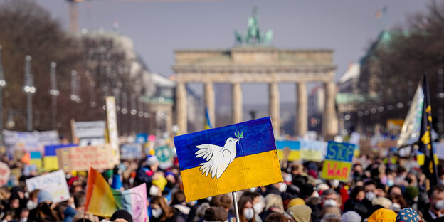 Friedensdemo in Berlin. Eine Demonstrantin hält vor dem Brandenburger Tor ein Schild mit der ukrainischen Flagge und einer weißen Friedenstaube hoch.