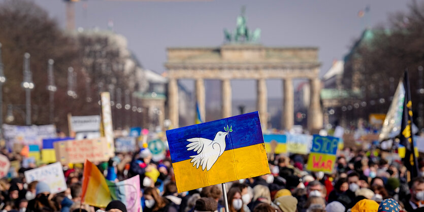 Friedensdemo in Berlin. Eine Demonstrantin hält vor dem Brandenburger Tor ein Schild mit der ukrainischen Flagge und einer weißen Friedenstaube hoch.