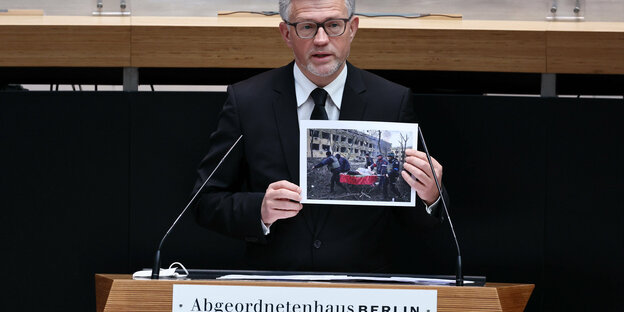 Das Bild zeigt den ukrainischen Botschafter Andrij Melnik mit einem Kriegsfoto in den Händen am Rednerpult des Abgeordnetenhauses.