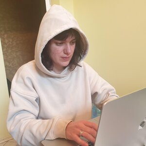 Eine Frau im hellen Kapuzenpulli mit Kapuze über dem Kopf vor einem aufgeklappten Laptop:Lizza Smith Eine Frau im hellen Kapuzenpulli mit Kapuze über dem Kopf vor einem aufgeklappten Laptop:Lizza Smith