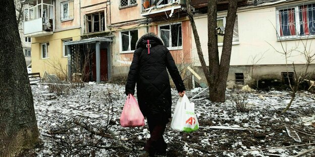 Eine Frau geht mit Tüten zu einem zerbombten Haus
