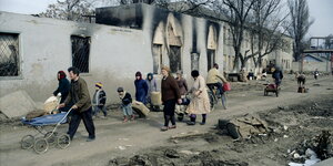 Menschen ziehen mit Koffern und Kinderwägen und Habseligkeiten vor zerschossenen Häusern auf einem Weg