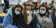 Drei Frauen mit Mundschutzmasken sitzen in Decken gehüllt in einer LAgerhalle