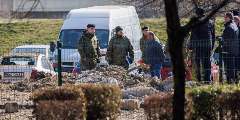 Mehrere Männer in Uniform stehen hinter hecken. Sie blicken auf den Boden, wo das FLugobjekt eingeschlagen ist. Hinter ihnen ein weißer Van.