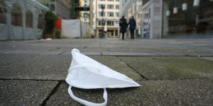 Eine FFP2-Maske liegt auf dem Weg in einer bayrischen Innenstadt. Es ist Tag und Im Hintergrund laufen zwei Personen.