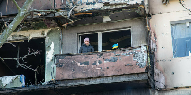 Bewohnerin eines Plattenbaus auf dem Balkon - mit ukrainischer Flagge - ihrer ausgebrannten Wohnung