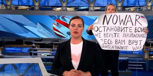 Hinter einer Nachrichtensprecherin in einem Fernsehstudio steht eine Person, die ein Plakat in die Kamera hält