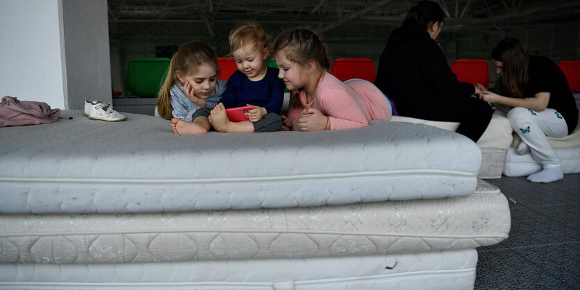 3 kleine Kinder schauen auf den Bildschirm eines roten Iphones, sie liegen auf aufgestapelten Matratzen