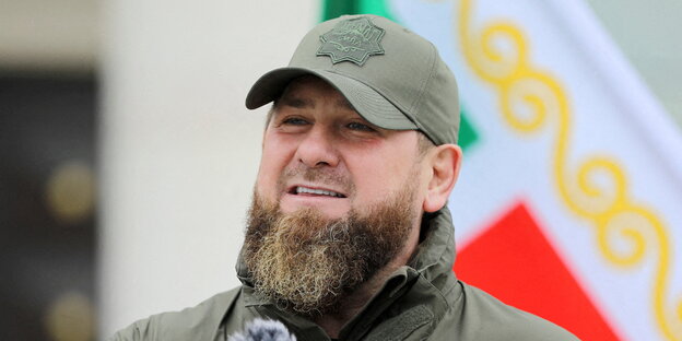 Kadyrow trägt ein Cap und Bart und steht vor einer Fahne