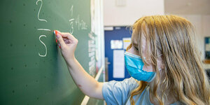 Ein Mädchen, das eine Mundschutzmaske trägt, schreibt Zahlen an eine Schultafel