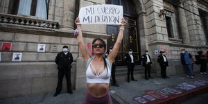 Eine junge Frau in Bikini-Top und kurzer Hose hält ein Schild hoch auf dem steht "Mein Körper, meine Entscheidung"; hinter ihr stehen Sicherheitsmänner.