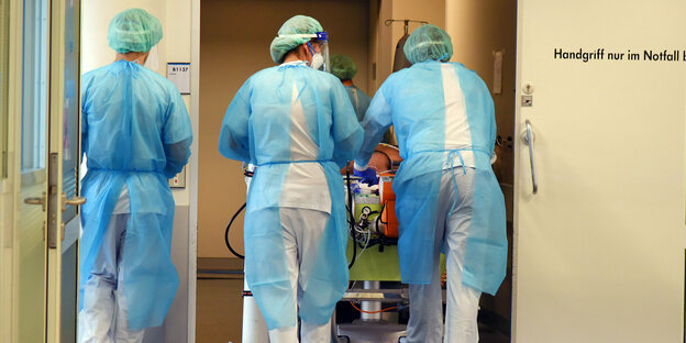 Drei Pflegende in Schutzkleidung fahren einen Patienten im Bett durch ein Krankenhaus in Leipzig.