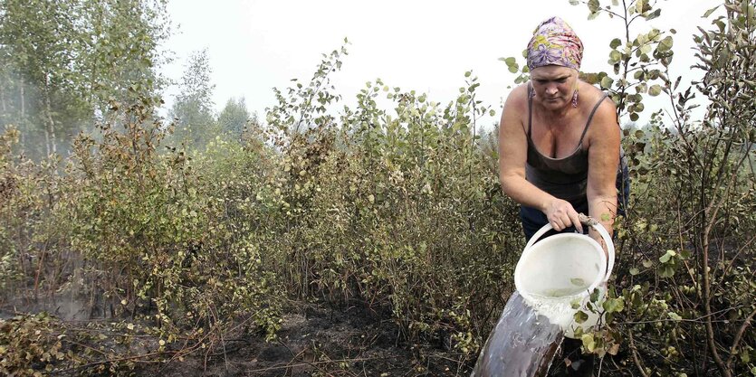 Eine Frau schüttet Wasser aus einem Plastikeimer auf eine dampfende Moorlandschaft. Um sie herum stehen mannshohes Gräser.