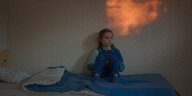 Nelly (Joséphine Sanz) sitzt auf einem Kinderbett, rötliches Licht auf der Wand über ihr.