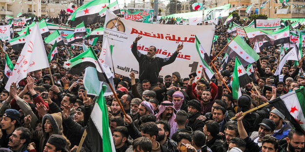 Ein lachender kleiner Junge hält eine syrische Revolutionsfahne, inmitten einer Gruppe von ebenfalls die Revolutionsfahne, die drei statt zwei Sterne abbildet, schwingenden Menschen