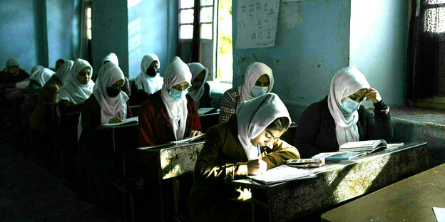 In einem wenig beleuchteten Raum sitzen junge Mädchen mit Kopftuch und Maske and Schulbänken und blicken konzentriert auf ihre Unterlagen.
