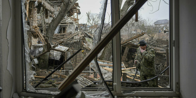 Durch ein kaputtes Fenster sieht man draußen einen Soldaten und viele Trümmer