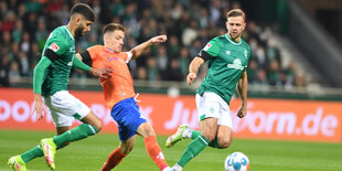 Ein Fußballspieler in gelb-blauem Trikots kämpft mit zwei Fußballspieler in grün-weißen Trikot rechts und links von iohm