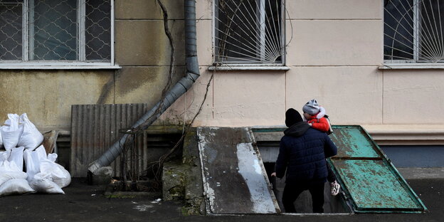Ein Mann bringt seine Tochter in einem Keller, daneben Sandsäcke vor einem Haus mit Rauchspuren