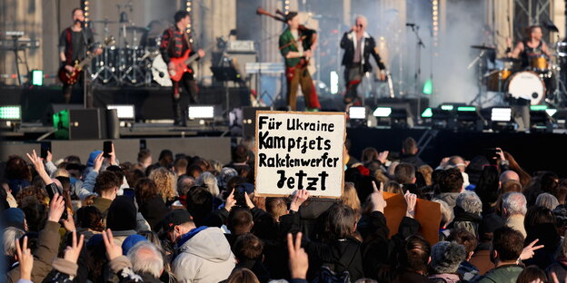 Kampfjets und Raketenwerfen für die Ukraine steht auf einem Plakat vor der Bühne, in der Unschärfe Sänger vor dem Brandenburger Tor