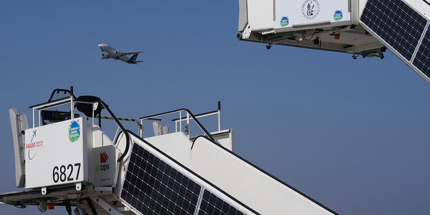 zwei Fluggastbrücken mit Solarzuellen, dazwischen ein Flugzeug am Himmel