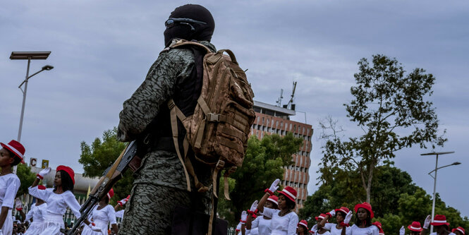 Ein Mann in Tarnkleidung Maske steht am Rande einer Parade von Mädchen in weißen Kleidern und roten Hüten. Der Soldat ist mit einem Sturmgewehr bewaffnet