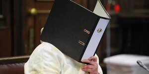 Eine Frau im Gerichtssaal hält einen Aktenordner vor den Kopf, um ihr Gesicht zu verbergen