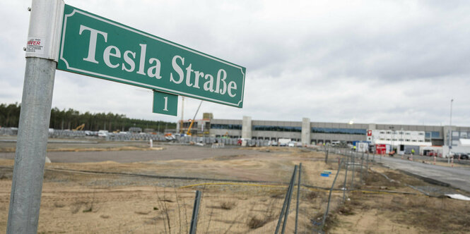 Das Bild zeigt im Vordergrund ein Straßenschild namens "Tesla-Straße", im Hintergrrund die jetzt eröffnete Tesla-Fabrik in Gründheide.