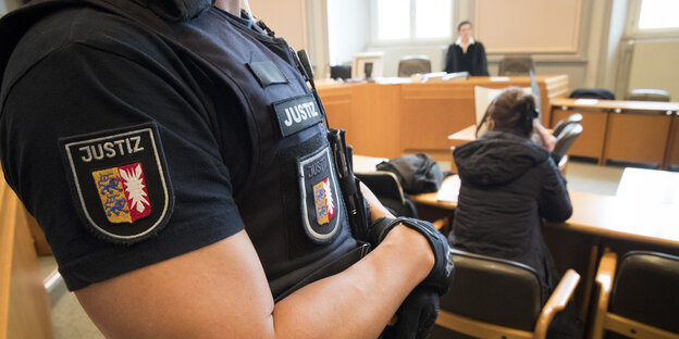 Ein Justizbeamter steht im Gerichtssaal, im Hintergrund des Bildes findet ein Prozess statt