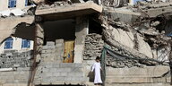Ein junge in weißen Gewand steht vor einem zerstörten sandfarbenen Gebäude.