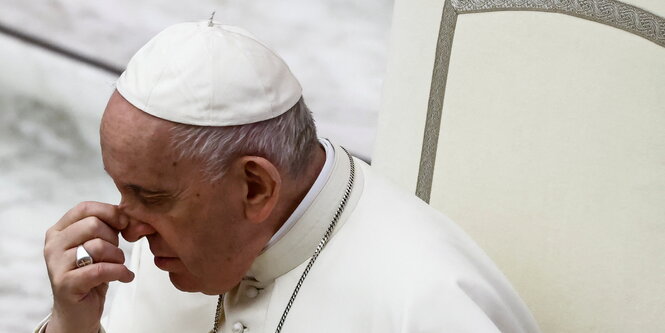 Papst Franziskus hält sich eine Hand vors Gesicht, er sitzt auf einem weißen Stuhl