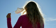 Eine junge Frau mit langem Haar von hinten, nimmt den Mundschutz ab