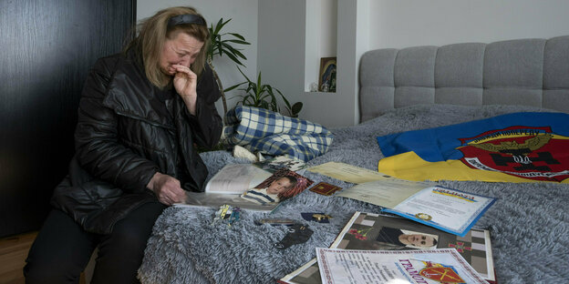 Weinende Mutter sitzt auf einem Bett, auf dem Urkunden und Dokumente ihres Sohnes und eine ukrainsiche Flagge liegen