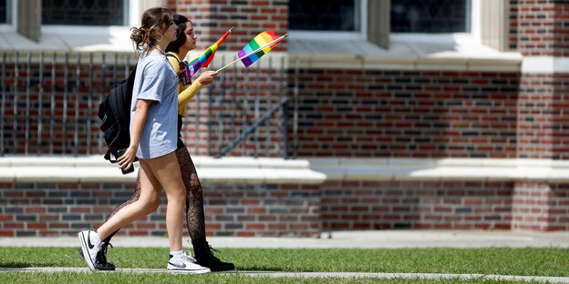 2 junge Frauen in Shorts gehen mit Regenbogenfahnen vor einem Gebäude