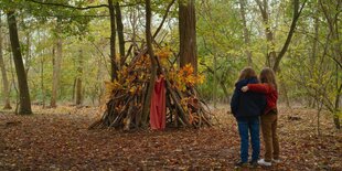 Zwei kleine Mädchen im Wald vor einer selbstgebauten Hütte aus Ästen