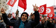 Mehrere Männer mit tunesischen Fahnen demonstrieren, der Mann in der Mitte des Bildes macht ein Peace-Zeichen.
