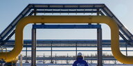 Eine blau angezogene Person mit dem Gazprom-Logo auf der Jacke geht in einer Schneelandschaft unter gelben und blauen Gaspipelines hindurch