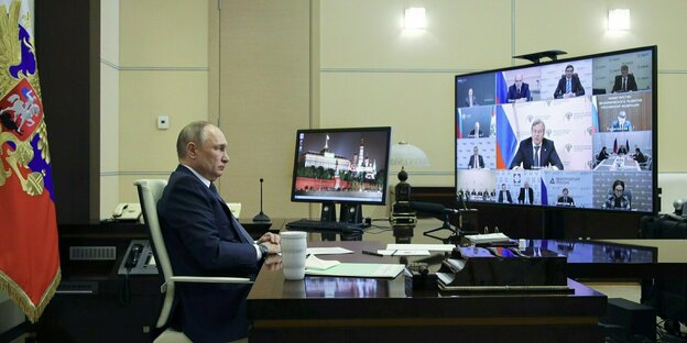 Wladimir Putin sitzt an einem Schreibtisch und schaut auf einen großen Bildschirm, der eine Videokonferenz zeigt