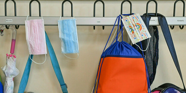 Masken hängen zusammen mit Taschen und Rucksäcken an Kleiderhaken in einem Klassenraum