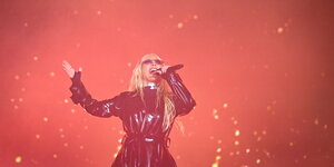 Eine schwarz gekleidete, blonde Frau singt vor einem rot beleuchteten Hintergrund