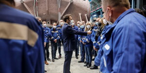 Macron umingt von Arbeitern in blauer Kleidung in der Werkshalle hält ein Mikro in der Hand