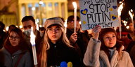 Zwei junge Frauen, und im Hintergrund weitere Menschen, halten Schilder hoch, auf denen sie Solidarität für die Ukraine bekunden, und brennende Kerzen.