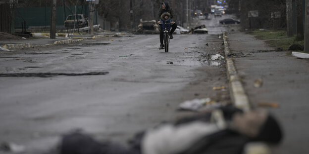 Ein Mann und ein Kind fahren auf einem Fahrrad, während die Leiche eines Zivilisten auf der Straße im ehemals russisch besetzten Kiewer Vorort Butscha liegt